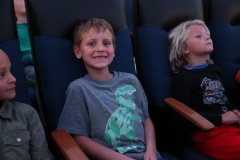 Three children sitting in movie theater seats, smiling at camera.