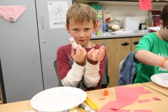 Young boy holding up a hand with two fingers extended in front of him, sitting at a table with craft materials and a plate