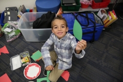 Boy holding a green leaf in front of him, surrounded by classroom supplies.