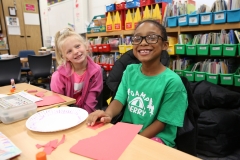 Two children sitting at a table in a classroom, working on crafts.