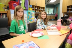 Two young girls sitting at a table in a classroom setting, engaged in an activity involving craft supplies and possibly a
