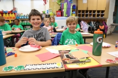 Two young boys sitting at a table in a classroom, smiling and looking at the camera. They are surrounded by school supplies
