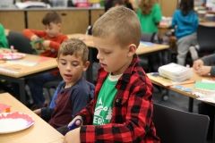 Two young boys sitting at desks in a classroom, working on craft projects.
