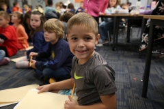 Young boy sitting at table in classroom, smiling at camera, holding pencil, surrounded by other children and adults.