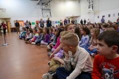 Children sitting on floor in gymnasium, watching event.