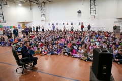 A large group of children gathered in a gymnasium for a reading event, listening to an adult sitting on a chair.