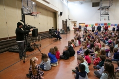 Children sitting on the floor in a gymnasium, watching a man speak at the front of the room.