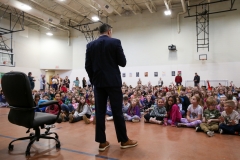 Man in suit standing among children in gymnasium.