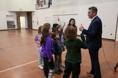 Man in suit giving thumbs up to children in gymnasium.