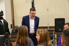Man in suit standing at basketball hoop, smiling, talking to children.
