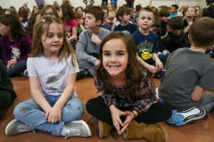 Children sitting on the floor in a classroom setting, smiling at the camera.