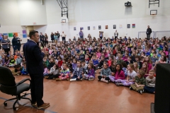 Man in suit standing among children seated on floor; he is speaking to them.