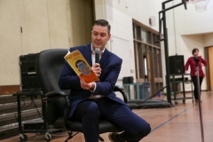 Man in suit holding book, speaking into microphone at event.