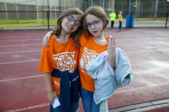 Two young girls posing together in front of an athletic track, both wearing glasses and orange shirts, one holding a blanket