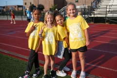 Four young girls posing together in yellow shirts at a track, smiling and giving peace signs.