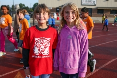 Two girls posing together at a sports event, both wearing red shirts with white lettering and one girl has a cat emblem on