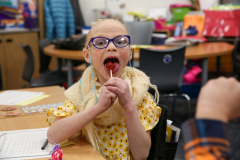 Girl in wheelchair licking a tootsie pop at her desk.