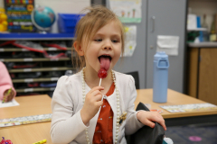 Young girl licking a tootsie pop in a classroom setting.