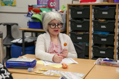 Old woman in glasses sitting at a table with a tootsie pop.
