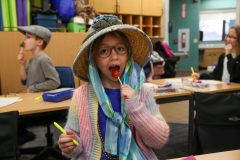 Girl in a room with tables, chairs, and other children. She is wearing glasses, a hat, and eating a lollipop.