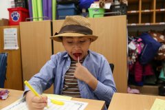 Child sitting at a desk in a classroom, licking a lollipop while holding a pencil and paper.