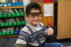 Young boy in glasses holding a lollipop, smiling at camera, sitting at a library desk.