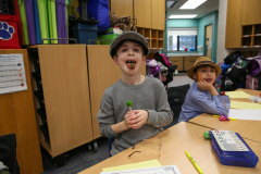 Two children in a classroom setting, one licking a tootsie pop, both wearing hats and sitting at a table with papers and
