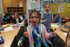 Two children in a classroom setting, one girl licking a tootsie pop, both smiling and enjoying their treats.