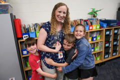 Woman in blue shirt hugging three children in a classroom setting.