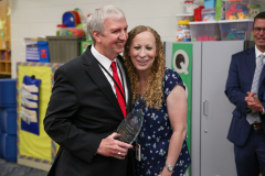 Two people posing together in a room filled with educational materials, one holding an award.