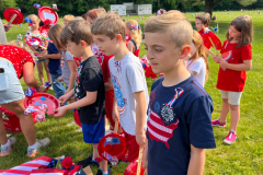 Children in patriotic attire, possibly at a parade or event, standing on grass and holding American flags.