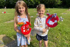 Two young girls posing in front of a grave decorated with red, white, and blue items.
