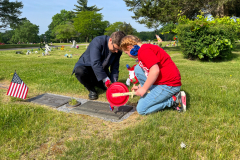 Two people at a grave site, one person is bending over to place a wreath on the headstone while another person stands nearby.