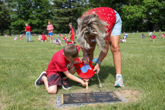 Woman in red shirt standing near grave marker. Child wearing red shirt kneeling down to place flowers on grave marker.