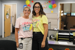 Two women posing in a room decorated for someone's first day of work, one wearing a pink shirt and the other a yellow shirt,
