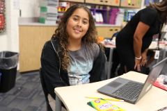 Young girl sitting at desk in classroom, smiling at camera.