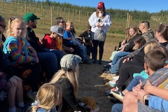 People gathered in a field, listening to someone speaking at an apple orchard event.