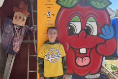 Young boy standing in front of a sign for an apple orchard.
