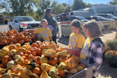 Three children standing in front of a table filled with pumpkins at an outdoor market.