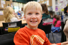 Young boy sitting at a table in a classroom, smiling at the camera.