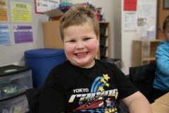 A young boy with a big smile sits at a desk in a classroom.