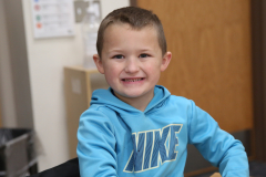 Young boy sitting at a table in a room, smiling towards the camera.