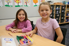 Two young girls sitting at a table in a classroom, smiling and looking towards the camera.