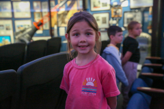 Little girl in pink shirt standing in front of movie theater seats.