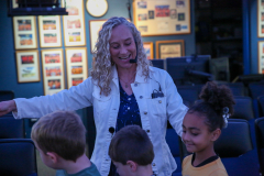 Woman in white jacket standing in front of children, gesturing with her hands while speaking into microphone.
