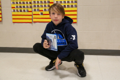 Person in blue hoodie holding a book, kneeling on one knee in front of a bulletin board.