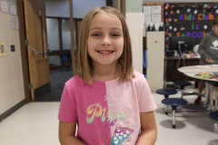 Little girl in pink shirt smiling at camera in school setting.