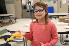 Little girl holding a slice of pizza in a school cafeteria.