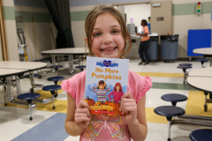 Girl holding a book in front of her face, smiling at the camera. She is standing in a room with tables and chairs, and there