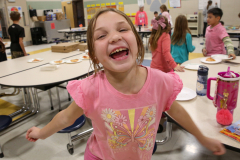 Girl in pink shirt laughing at camera in school cafeteria.